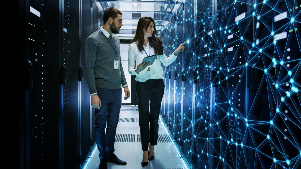 A man and woman stand in a futuristic server room, discussing data displayed on a glowing, networked digital interface - Managed IT Cloud Services