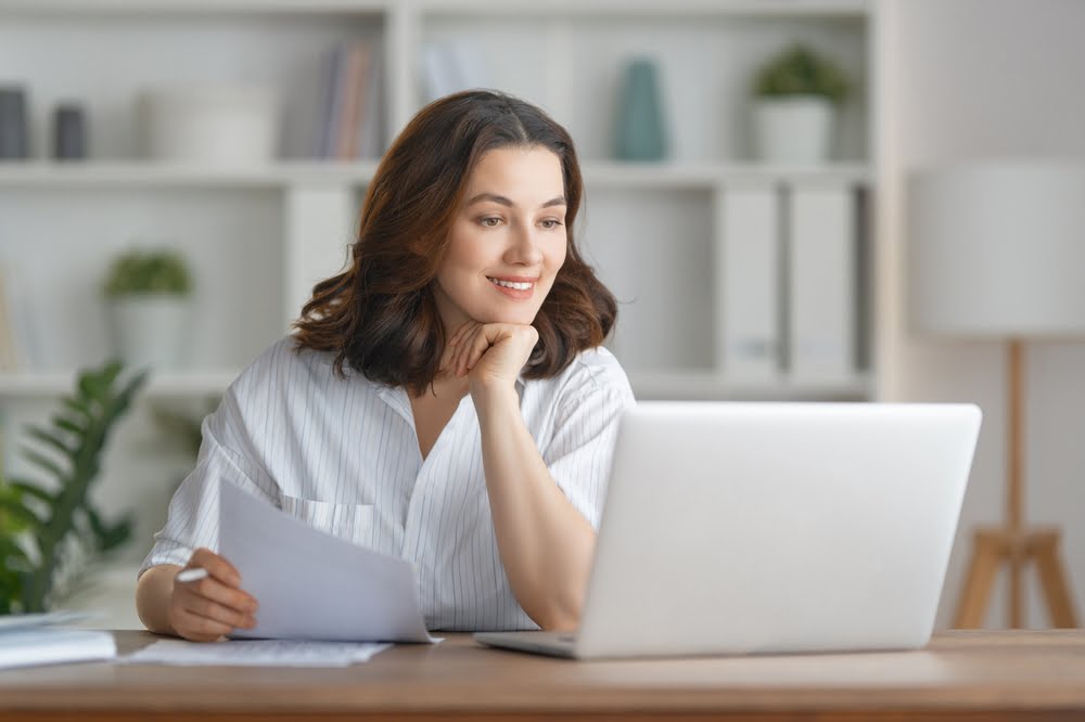 A smiling woman holding a paper works on her laptop at a wooden desk in a bright, organized home office - managed it services Riverside A smiling woman holding a paper works on her laptop at a wooden desk in a bright, organized home office - managed it services Riverside