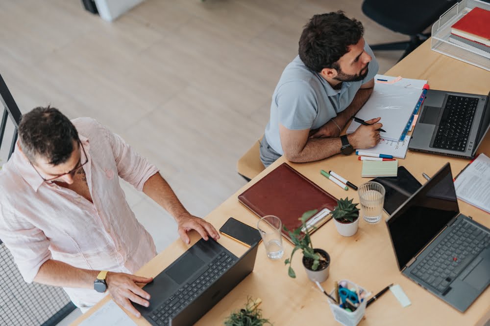 Two men collaborate at a shared desk with laptops, documents, and office supplies, viewed from above in a bright workspace - managed it services Rancho Cucamonga Two men collaborate at a shared desk with laptops, documents, and office supplies, viewed from above in a bright workspace - managed it services Rancho Cucamonga
