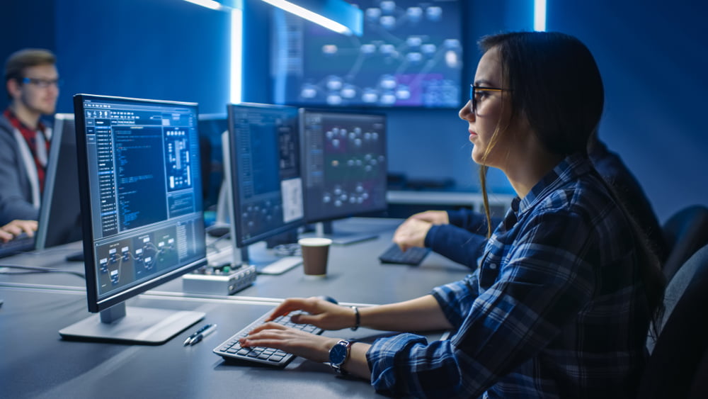 A woman in glasses working at a desk with multiple computer monitors displaying data and systems in a dimly lit, high-tech office environment - Managed IT Cloud Services