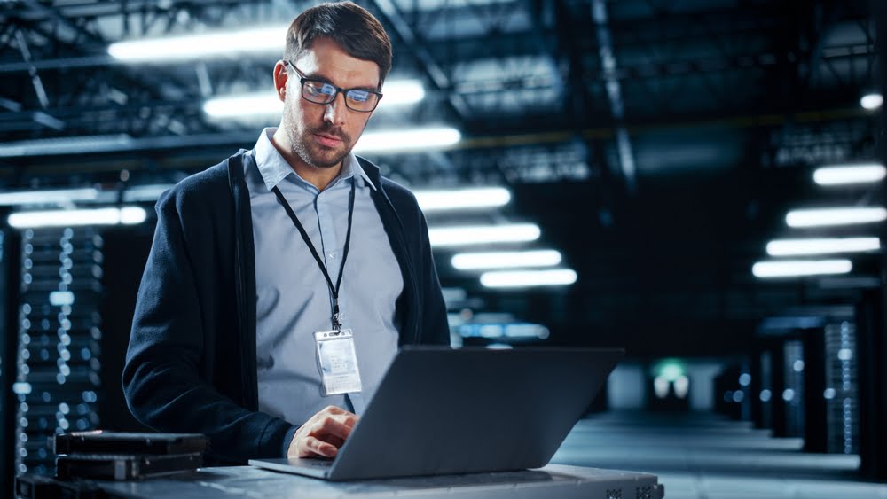 A man wearing glasses and an ID badge works on a laptop in a dimly lit server room with rows of illuminated servers in the background - Managed IT Cloud Services
