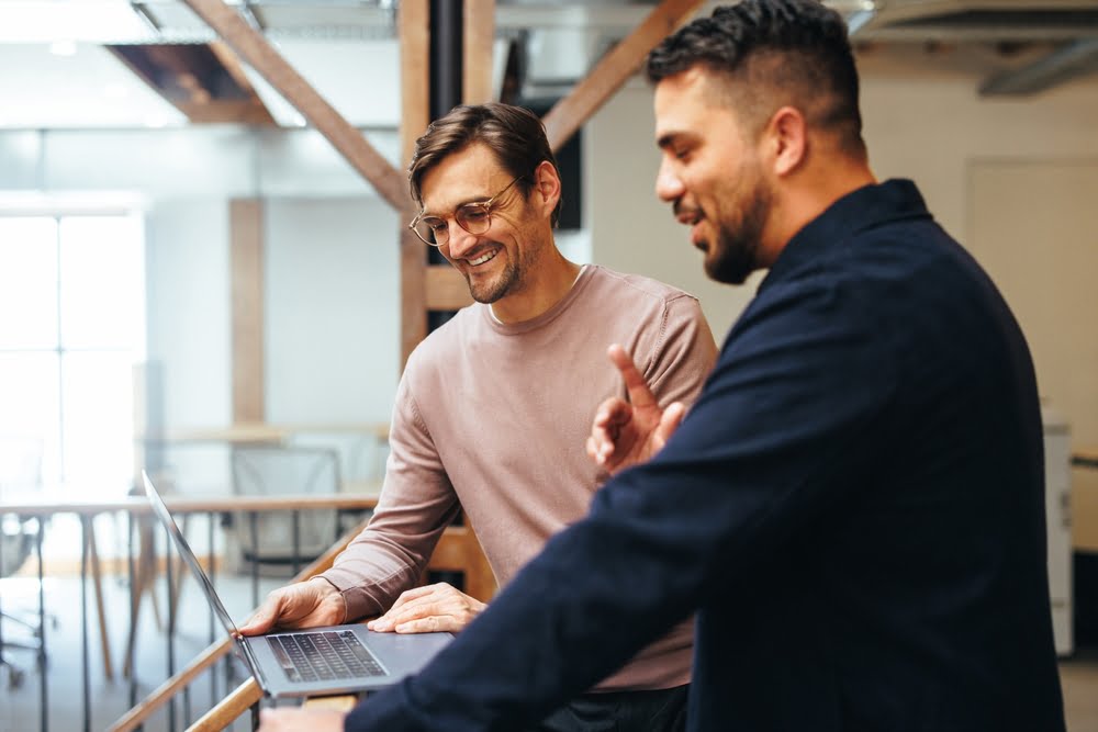Two men in a modern office setting are collaborating and smiling while using a laptop - managed it services temecula Two men in a modern office setting are collaborating and smiling while using a laptop - managed it services temecula