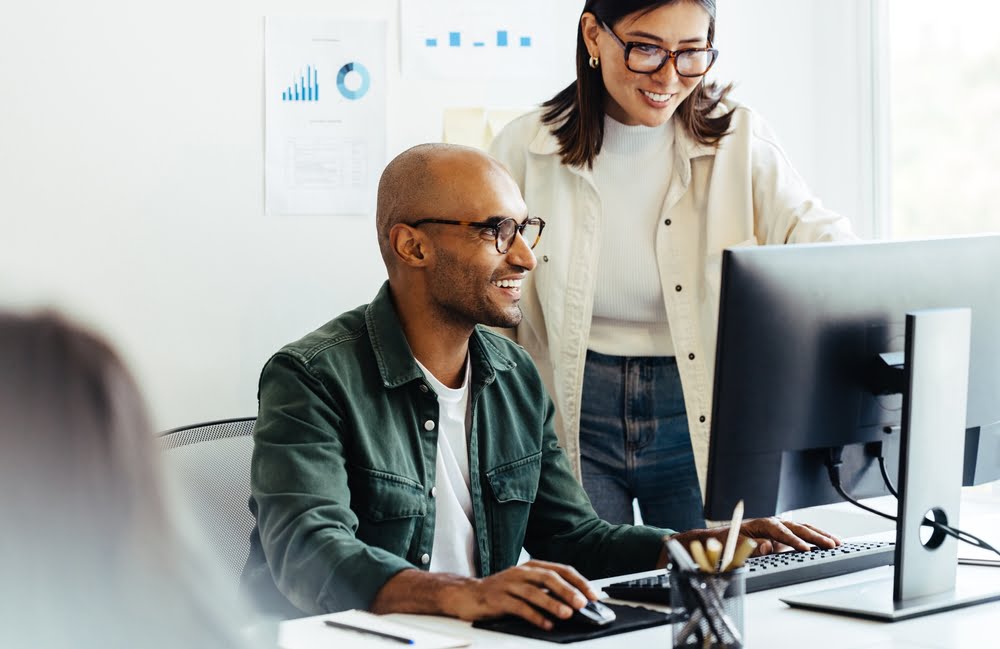 A man working on a computer smiles while a woman standing beside him looks at the screen in a modern office setting - managed it services Rancho Cucamonga A man working on a computer smiles while a woman standing beside him looks at the screen in a modern office setting - managed it services Rancho Cucamonga