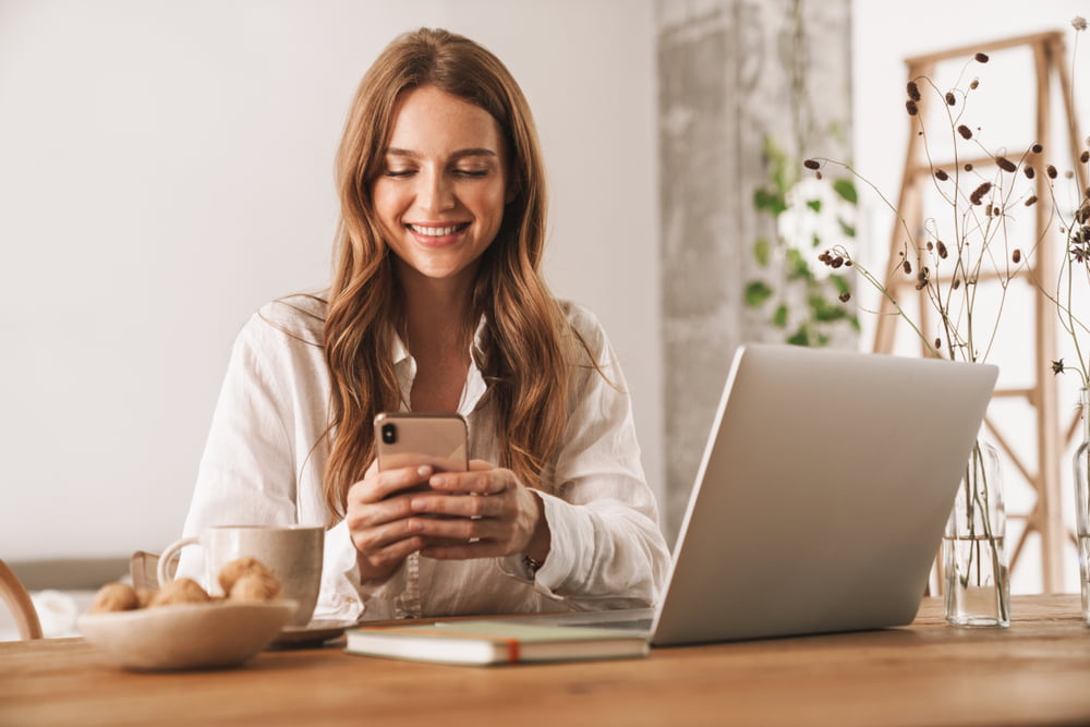 A smiling woman sits at a wooden table using her smartphone, with a laptop, notebook, coffee mug, and a bowl of snacks nearby - managed it services Rancho Cucamonga A smiling woman sits at a wooden table using her smartphone, with a laptop, notebook, coffee mug, and a bowl of snacks nearby - managed it services Rancho Cucamonga
