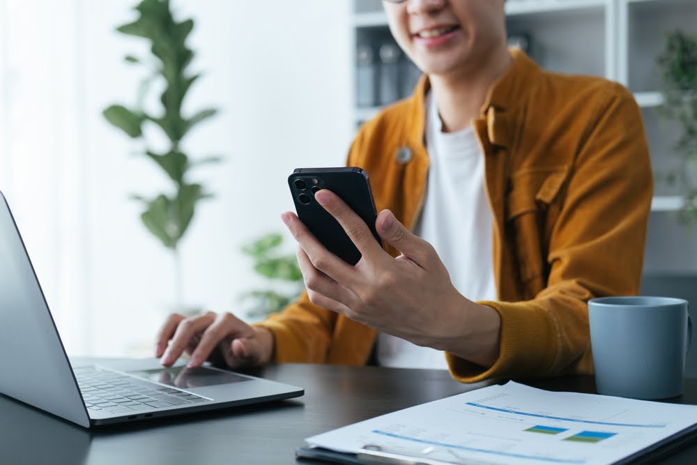A person smiling while holding a smartphone and working on a laptop at a desk with documents, a coffee mug, and plants in the background - Managed IT Services Corona A person smiling while holding a smartphone and working on a laptop at a desk with documents, a coffee mug, and plants in the background - Managed IT Services Corona