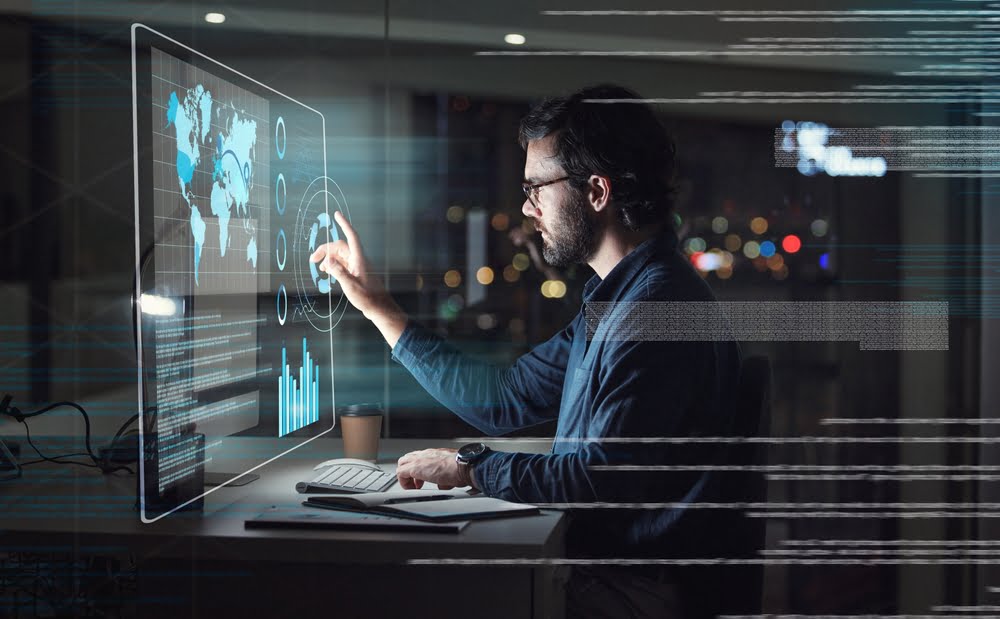 A man working at a desk interacts with a futuristic digital screen displaying data, maps, and graphs, in a dimly lit office with city lights in the background - Managed IT Services Corona A man working at a desk interacts with a futuristic digital screen displaying data, maps, and graphs, in a dimly lit office with city lights in the background - Managed IT Services Corona