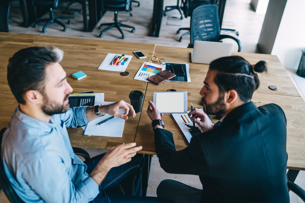Two men in a modern office are engaged in a discussion, with documents, a tablet, and colorful charts spread on the table between them - managed it services temecula Two men in a modern office are engaged in a discussion, with documents, a tablet, and colorful charts spread on the table between them - managed it services temecula