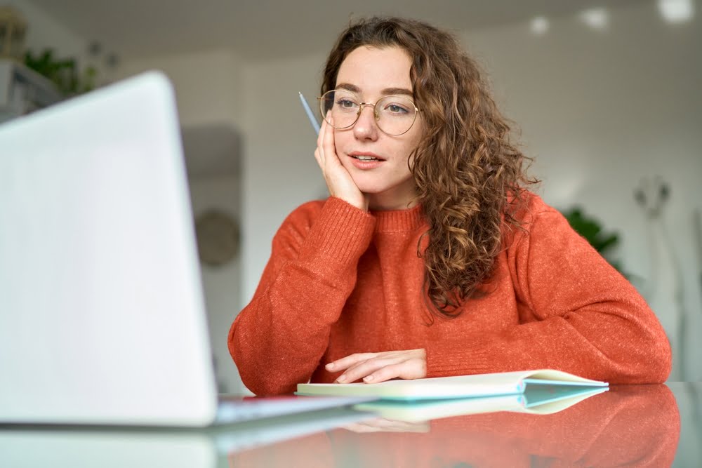 A young woman wearing glasses and an orange sweater looks at her laptop with interest, resting her head on her hand, with a notebook and pen on the table in a cozy setting - managed it services Rancho Cucamonga A young woman wearing glasses and an orange sweater looks at her laptop with interest, resting her head on her hand, with a notebook and pen on the table in a cozy setting - managed it services Rancho Cucamonga